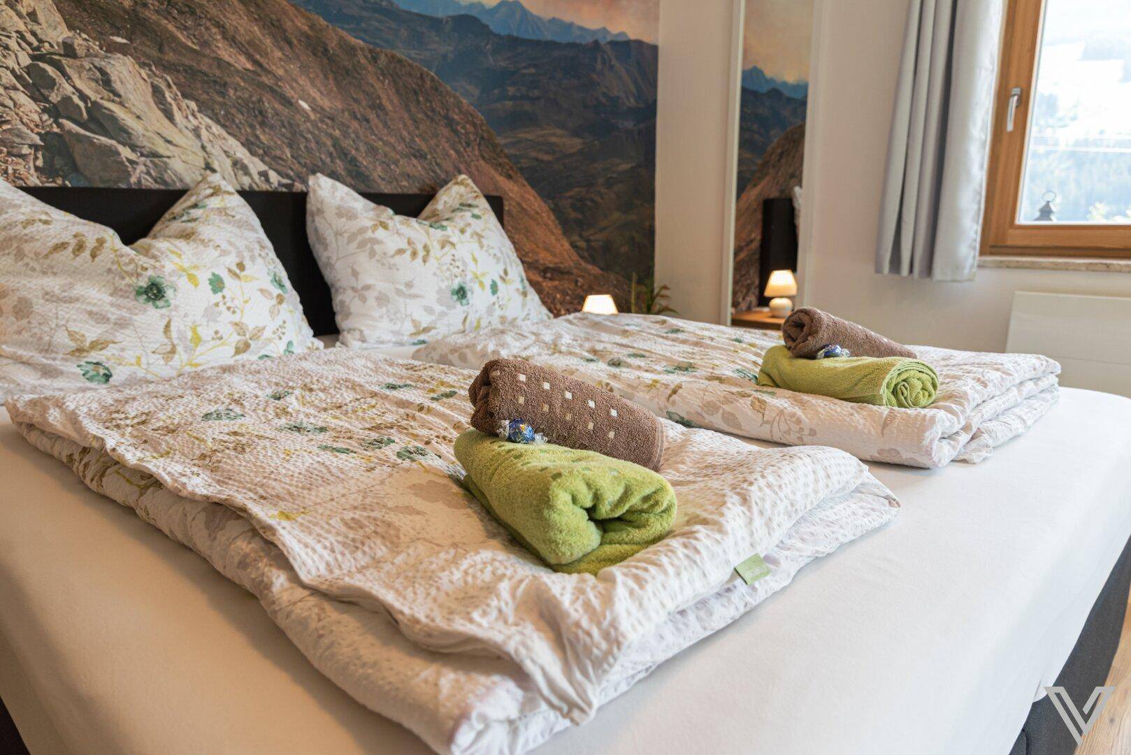 Gemütliches Doppelbett mit farbigen Handtüchern und Bergblick-Wandbild für eine entspannte Atmosphäre.