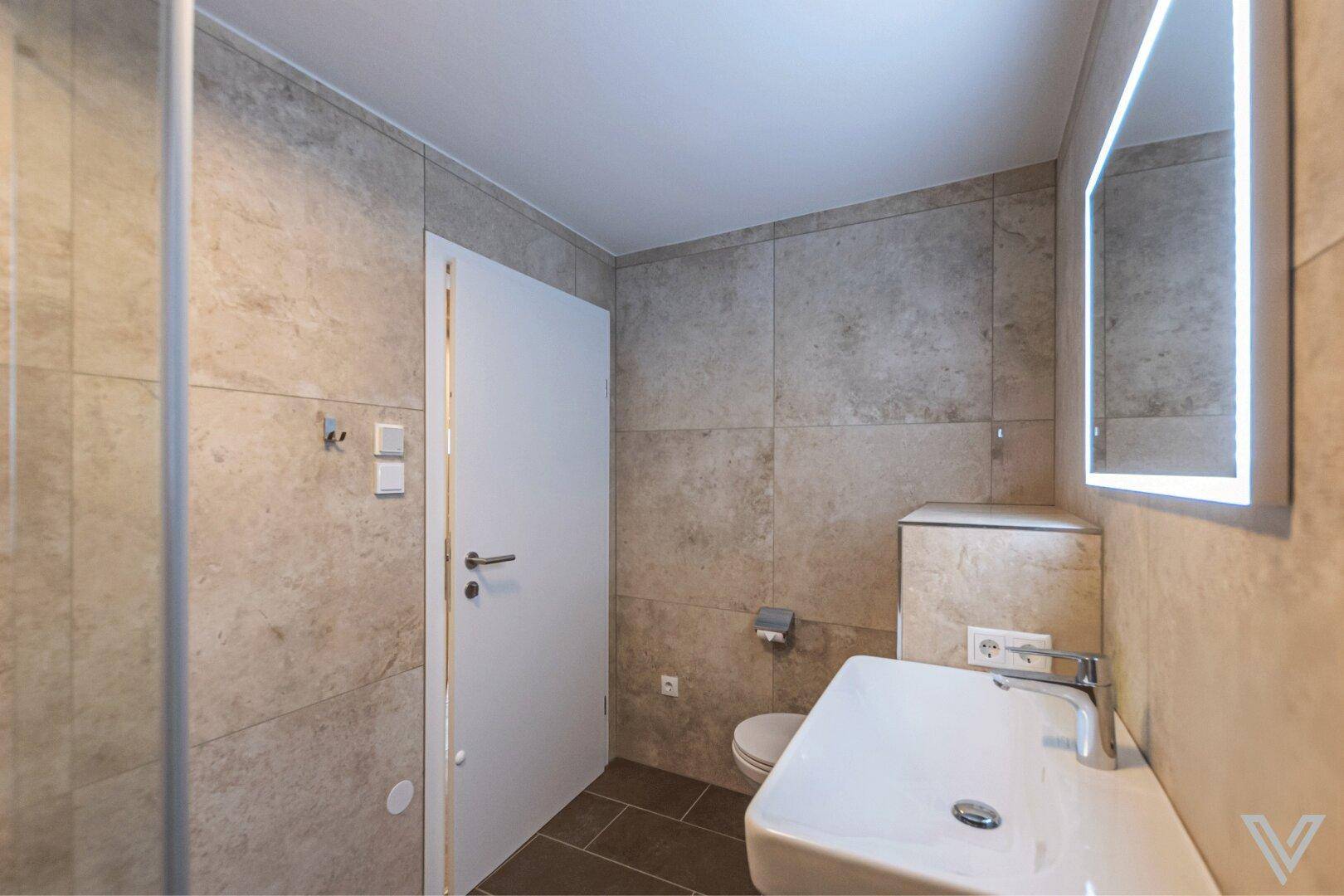 Modernes Badezimmer mit beigefarbenen Fliesen, beleuchtetem Spiegel und wandhängender Toilette.