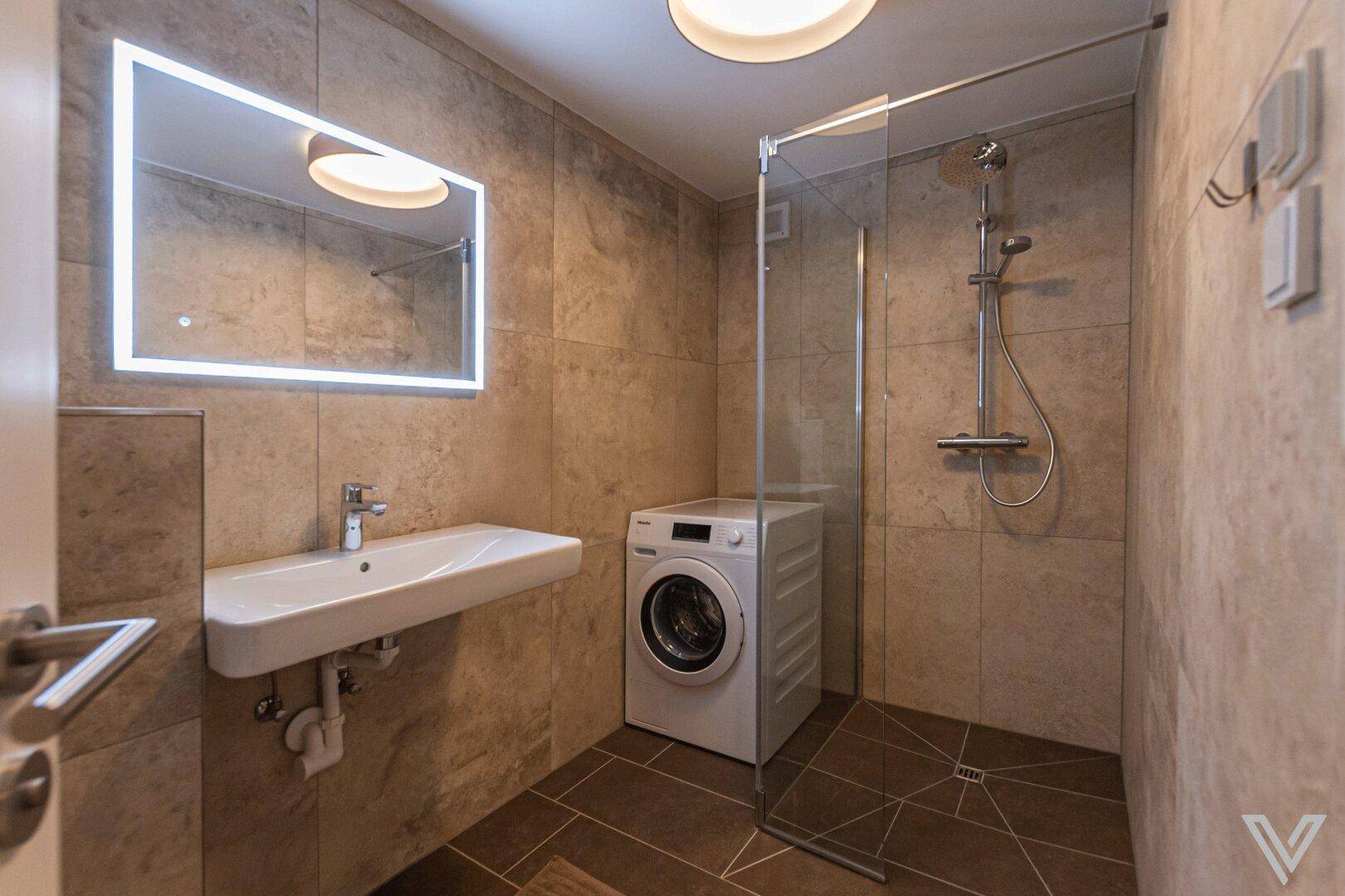 Helles Badezimmer mit Dusche, Waschmaschine und beleuchtetem Spiegel für Komfort.