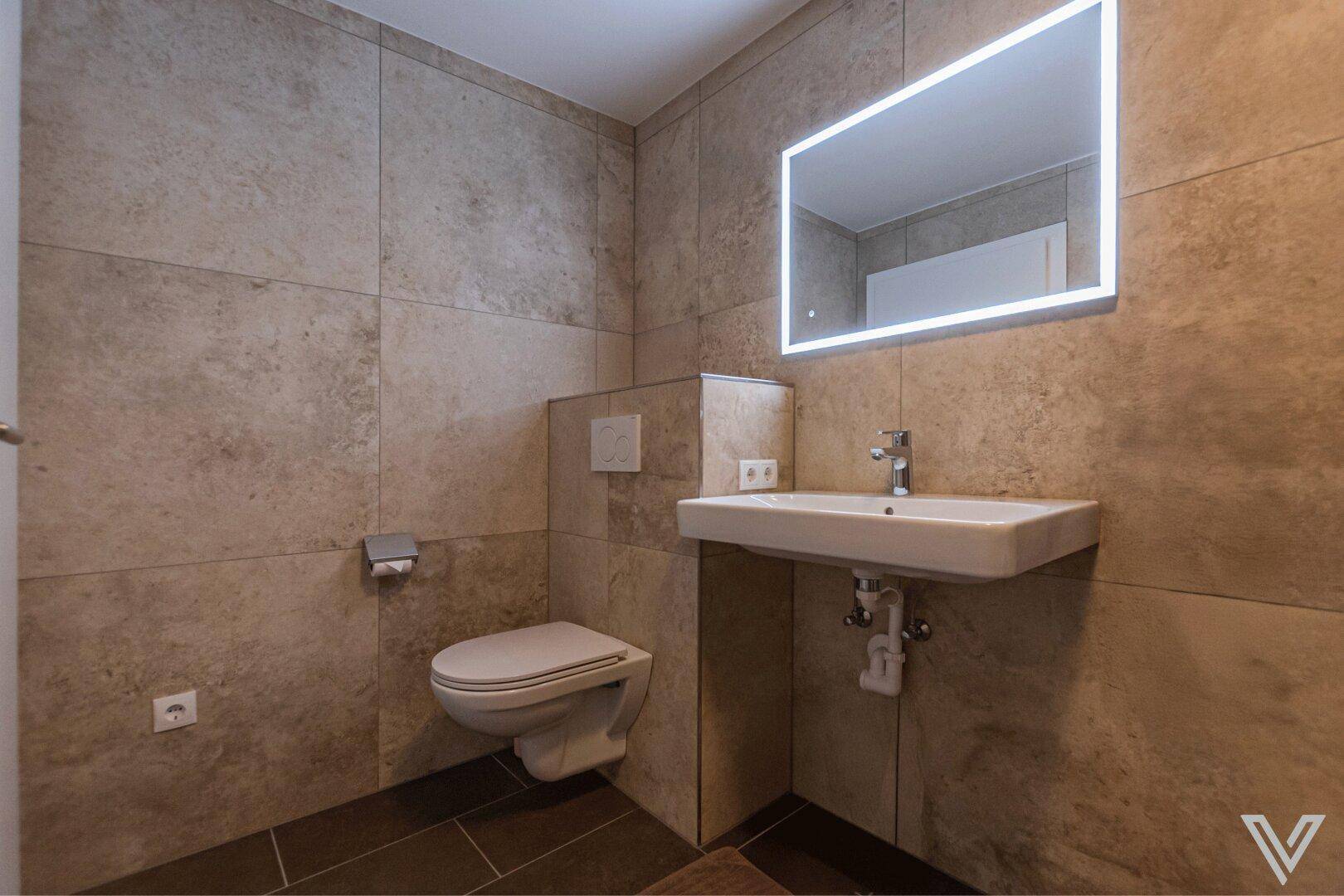 Modernes Badezimmer mit Waschbecken, Toilette und beleuchtetem Spiegel für eine angenehme Atmosphäre.