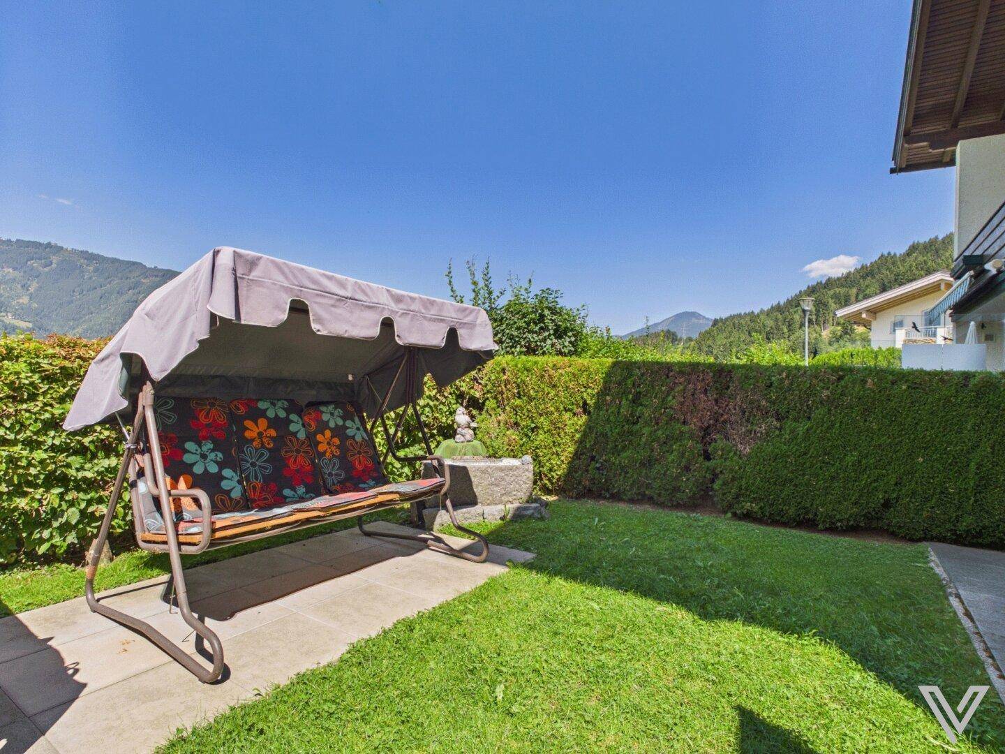 Gepflegter Gartenbereich mit Rasenfläche, Hecke und einer Hollywoodschaukel unter blauem Himmel.