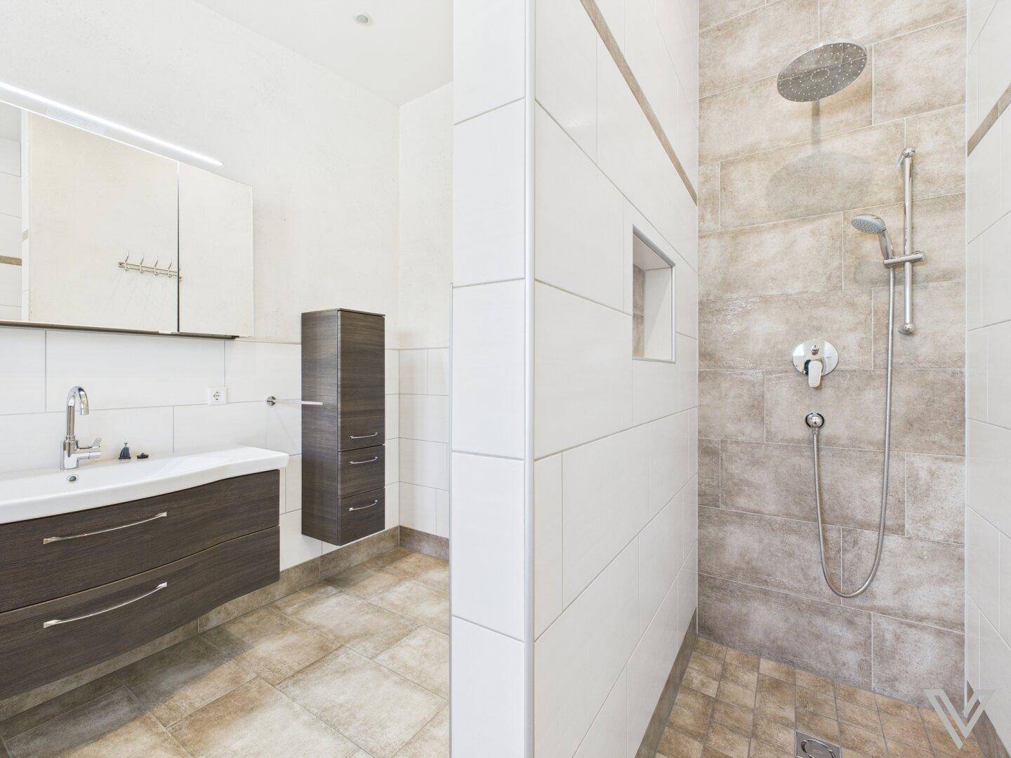 Modernes Badezimmer mit ebenerdiger Dusche und Regendusche, hellen Fliesen und dunklem Waschtisch.