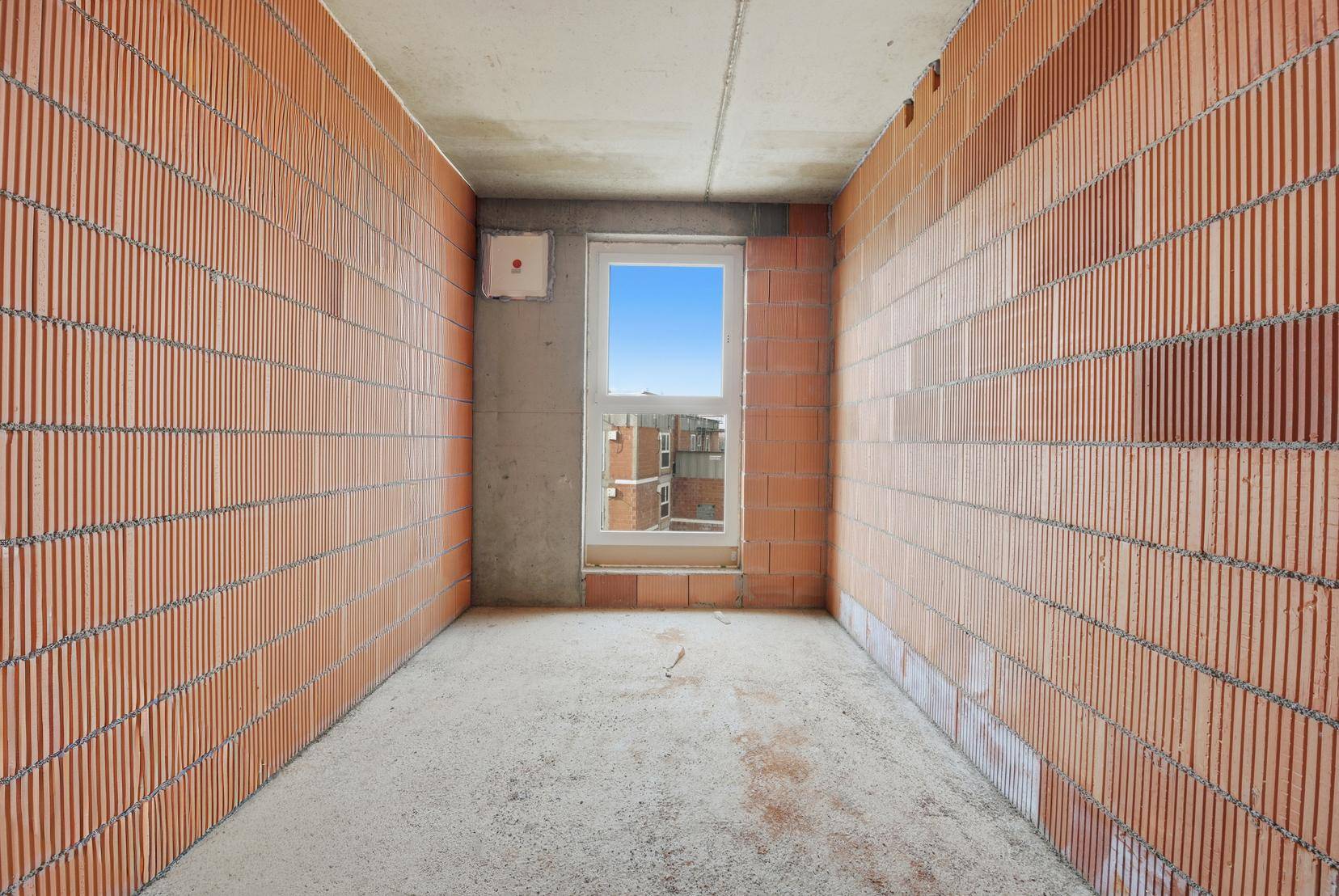 Schmaler Rohbau-Raum mit Ziegelwänden und einem einzelnen Fenster am Ende, das Tageslicht hereinlässt.