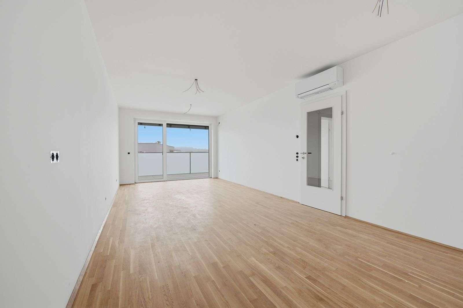 Unmöblierter Wohnbereich mit Holzboden, großen Fenstern und Zugang zum Balkon.