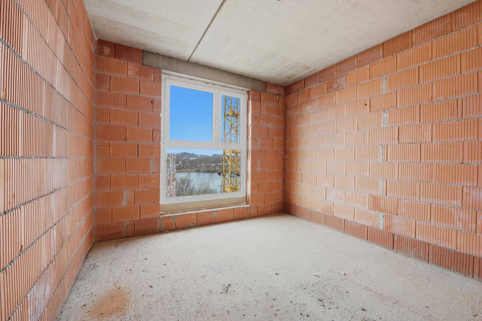 Unfertiger Raum mit Ziegelwänden und einem Fenster, das einen Blick auf den See und einen Kran freigibt.