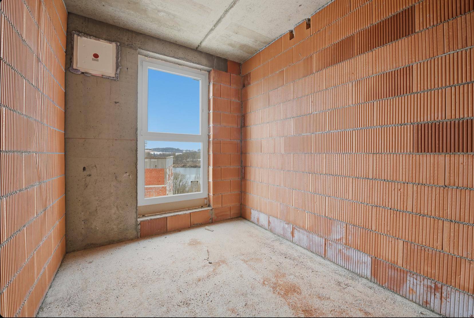 Unfertiges Zimmer mit Ziegelwänden und Betonboden, mit Blick auf die Baustelle und den See durch das Fenster.