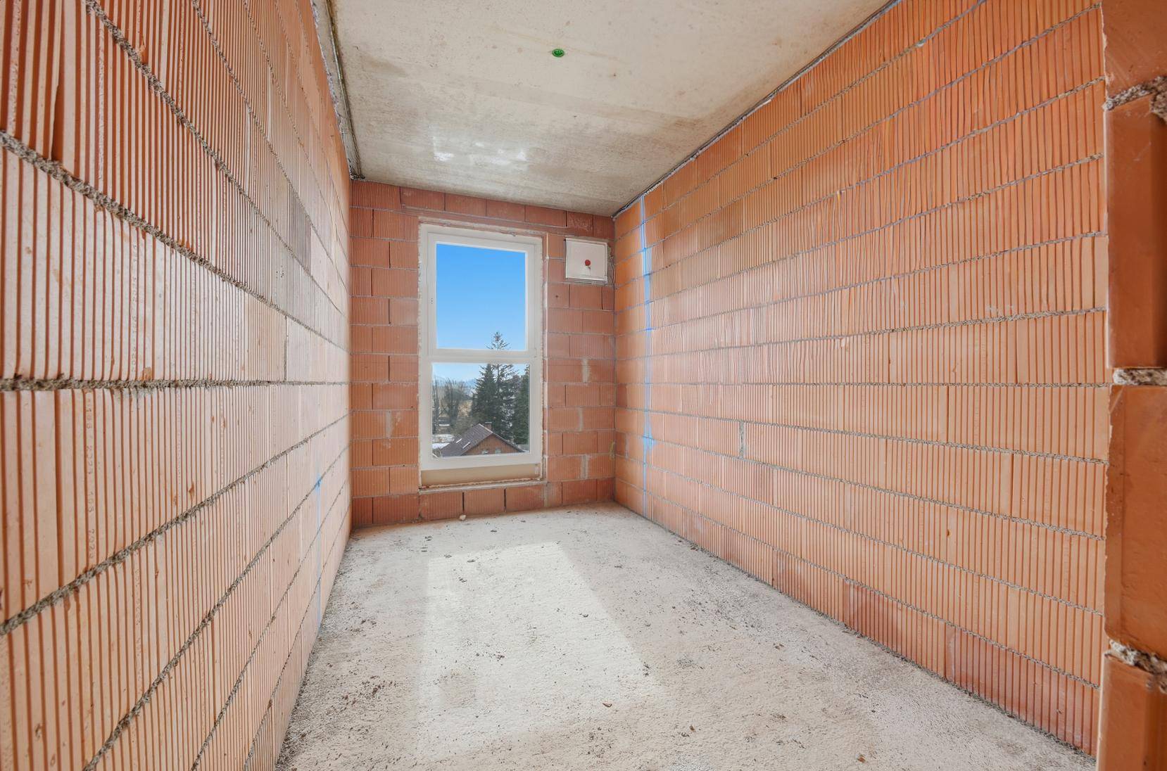 Unverputzter Rohbau mit Ziegelwänden und einem Fenster, das einen Blick ins Grüne bietet.