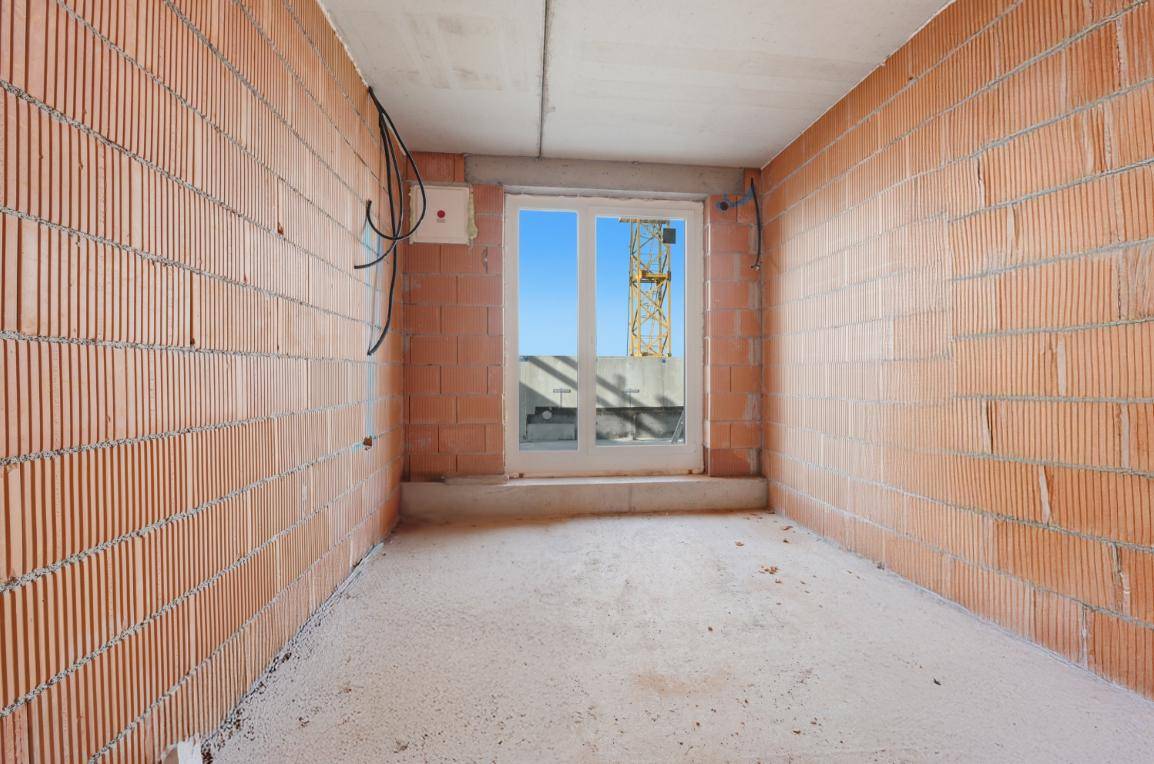 Unmöblierter Raum im Rohbau mit Ziegelwänden, Betonboden und Blick auf die Baustelle.
