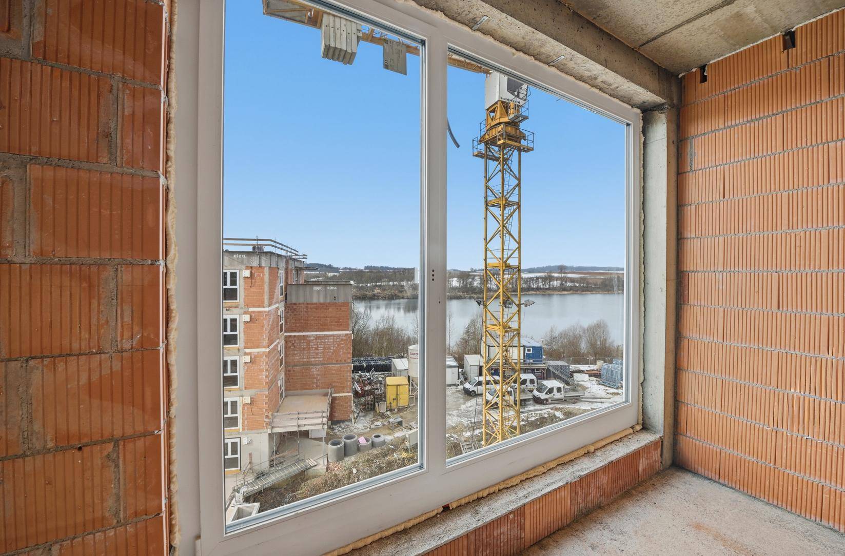 Unfertiger Innenraum mit großen Fenstern, die einen weiten Blick auf einen See und eine Baustelle bieten.