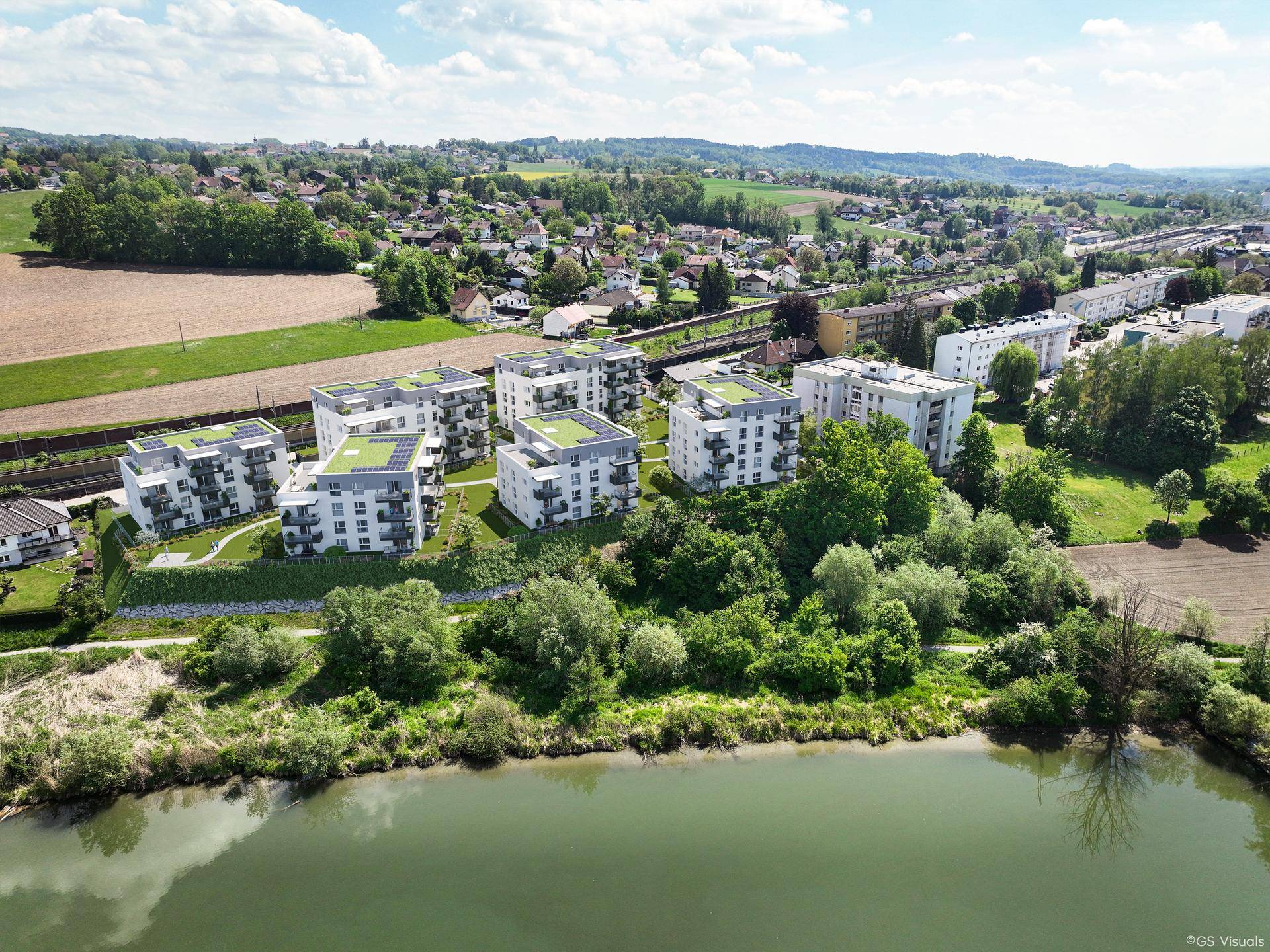 Luftaufnahme einer modernen Wohnanlage mit Flachdächern und Solaranlagen direkt am Wasser, umgeben von viel Grün.