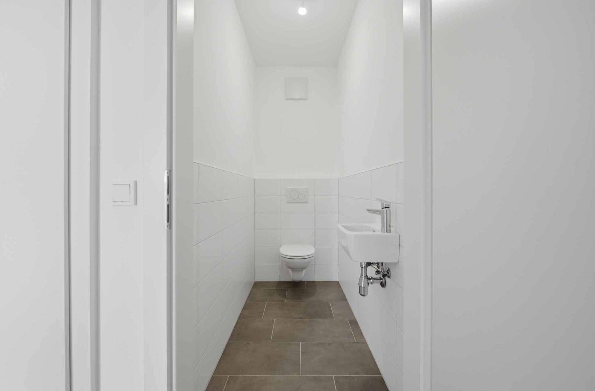 Modernes Gäste-WC mit weißer Wandfliese, wandhängender Toilette und kleinem Waschbecken.