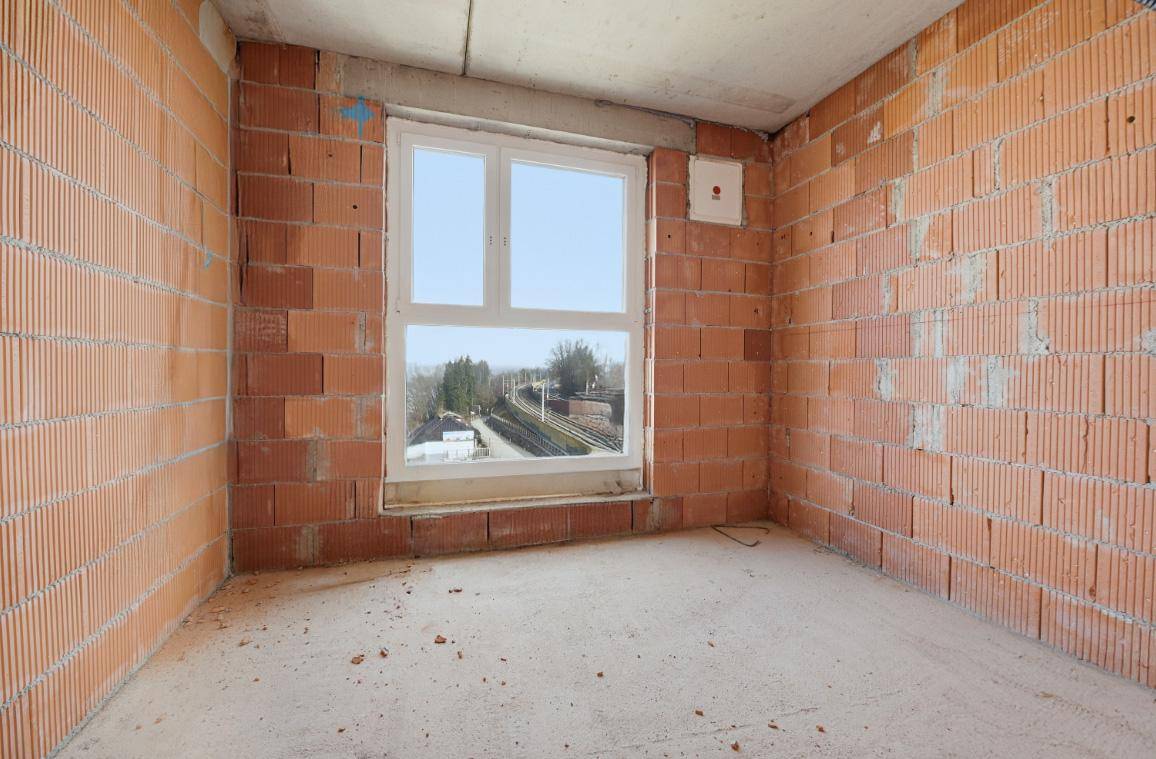 Rohbauzimmer mit Ziegelwänden, Betonboden und Fensterblick auf Bahngleise.