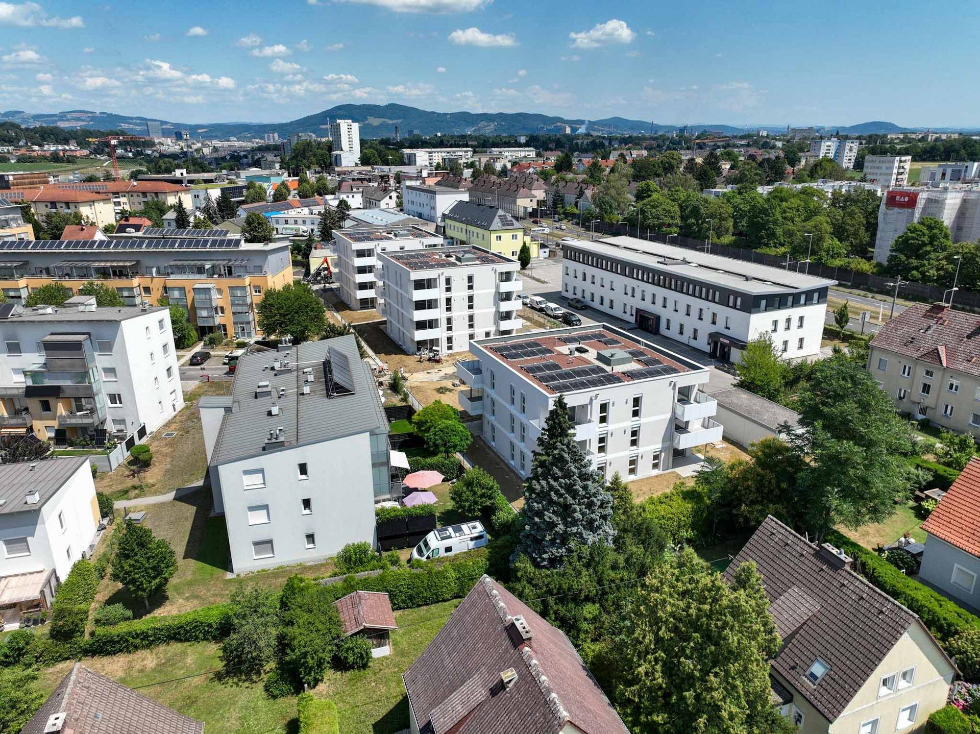 Weitläufige Luftaufnahme einer Wohnsiedlung mit neuen Gebäuden und älteren Häusern in grüner Umgebung.