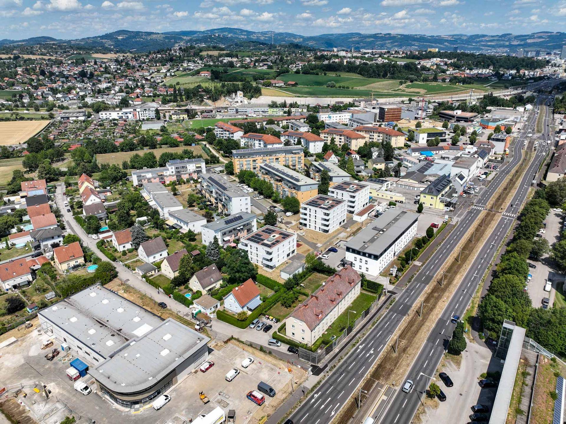 Weitläufige Luftaufnahme einer Stadtlandschaft mit Wohngebäuden, Straßen und grünen Hügeln im Hintergrund.