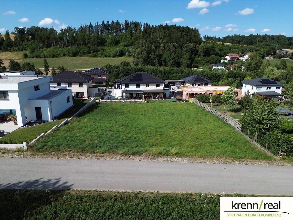 Blick auf ein grünes Grundstück mit modernen Häusern im Hintergrund und bewaldeten Hügeln unter blauem Himmel.
