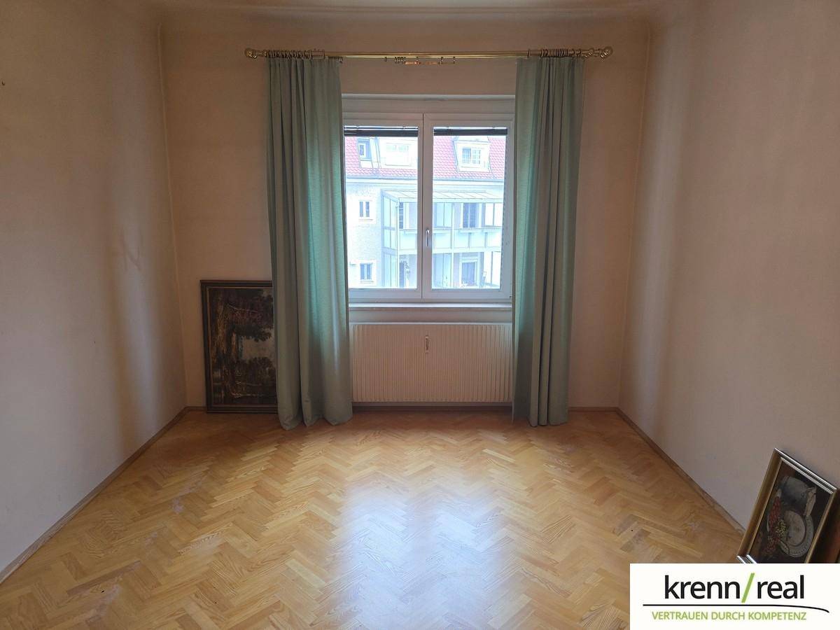 Leeres Zimmer mit Parkettboden, Fenster und hellen Vorhängen.