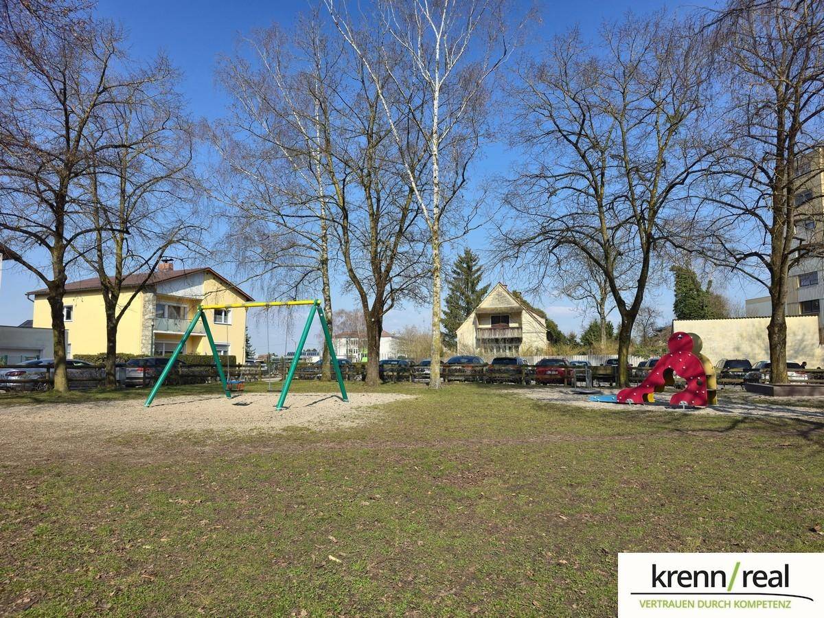 Kinderspielplatz mit Schaukeln und Klettergerüst, umgeben von Bäumen und Grünflächen.