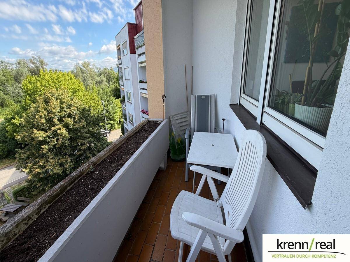 Schmaler Balkon mit Sitzgelegenheit, Blumenkasten und Ausblick auf Bäume und Nachbargebäude.