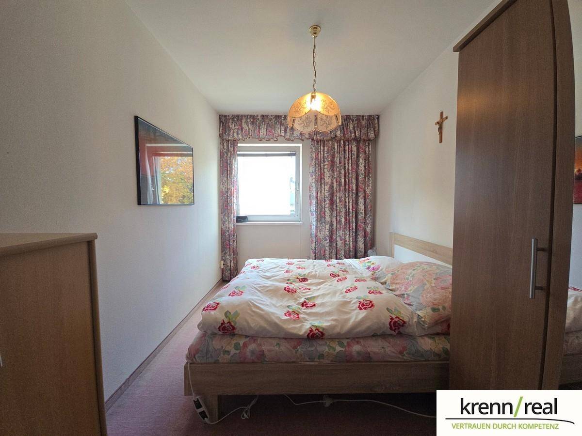 Schlafzimmer mit Doppelbett, Kleiderschrank und Fenster mit floralen Vorhängen.