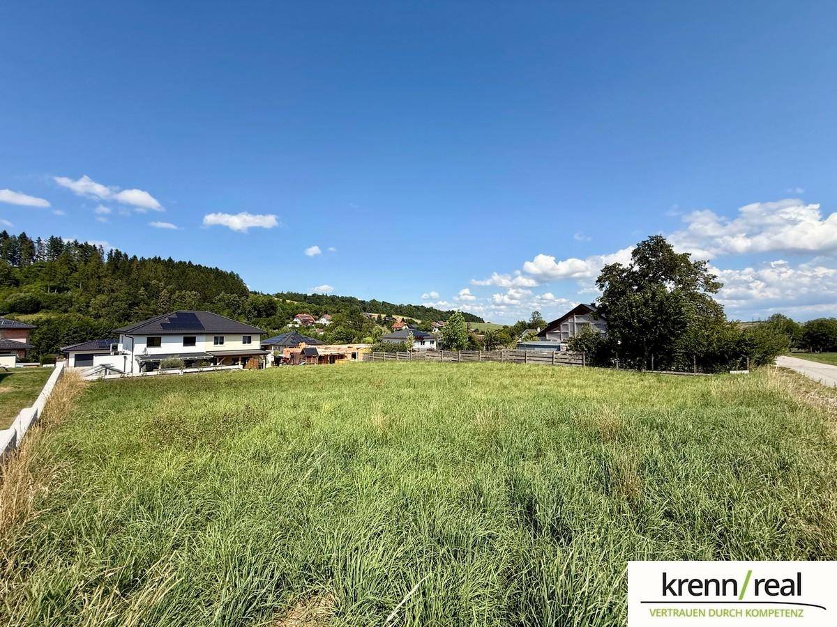 Grünes Baugrundstück mit Blick auf umliegende Wohnhäuser und eine ländliche Landschaft unter klarem Himmel.