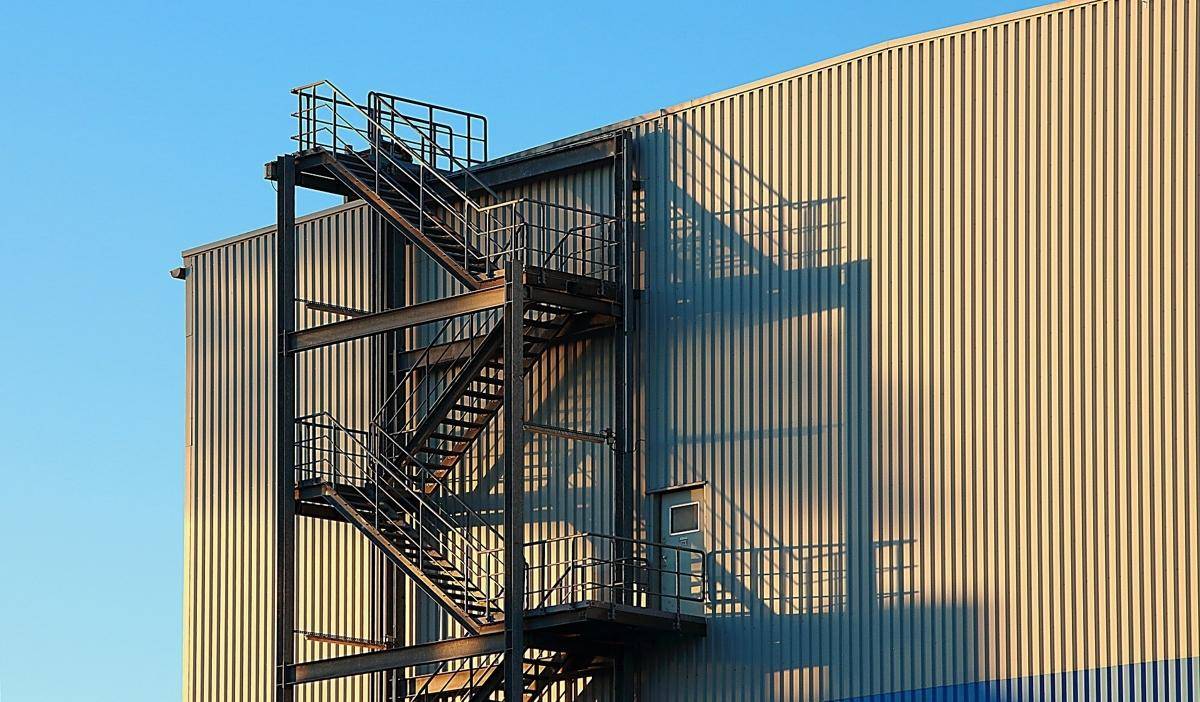 Außenansicht eines Gebäudes mit geriffelter Metallfassade und einer externen Feuertreppe unter blauem Himmel.