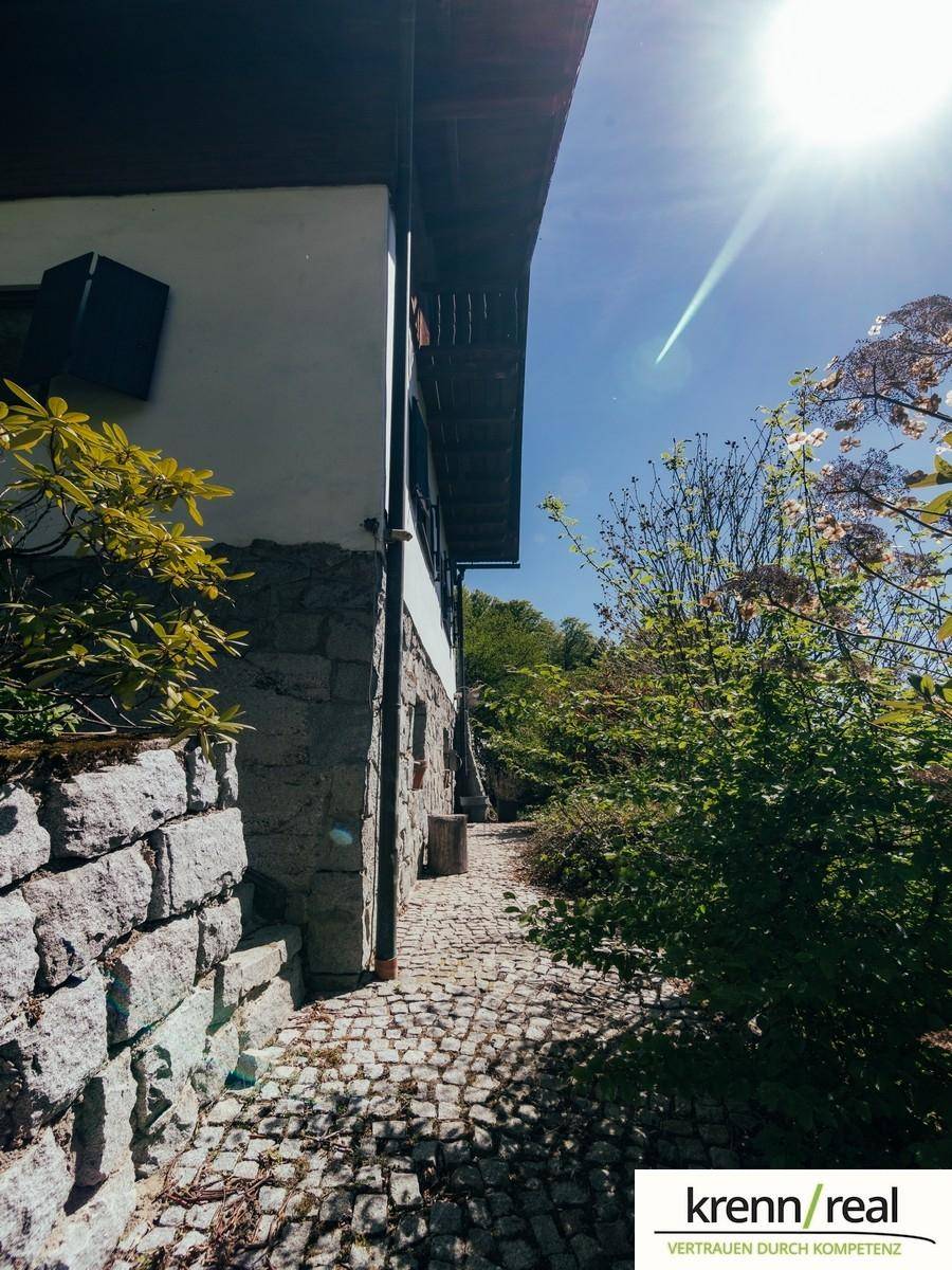 Kopfsteinpflasterweg entlang der Hauswand mit Steinmauer und üppiger Vegetation im Sonnenlicht.