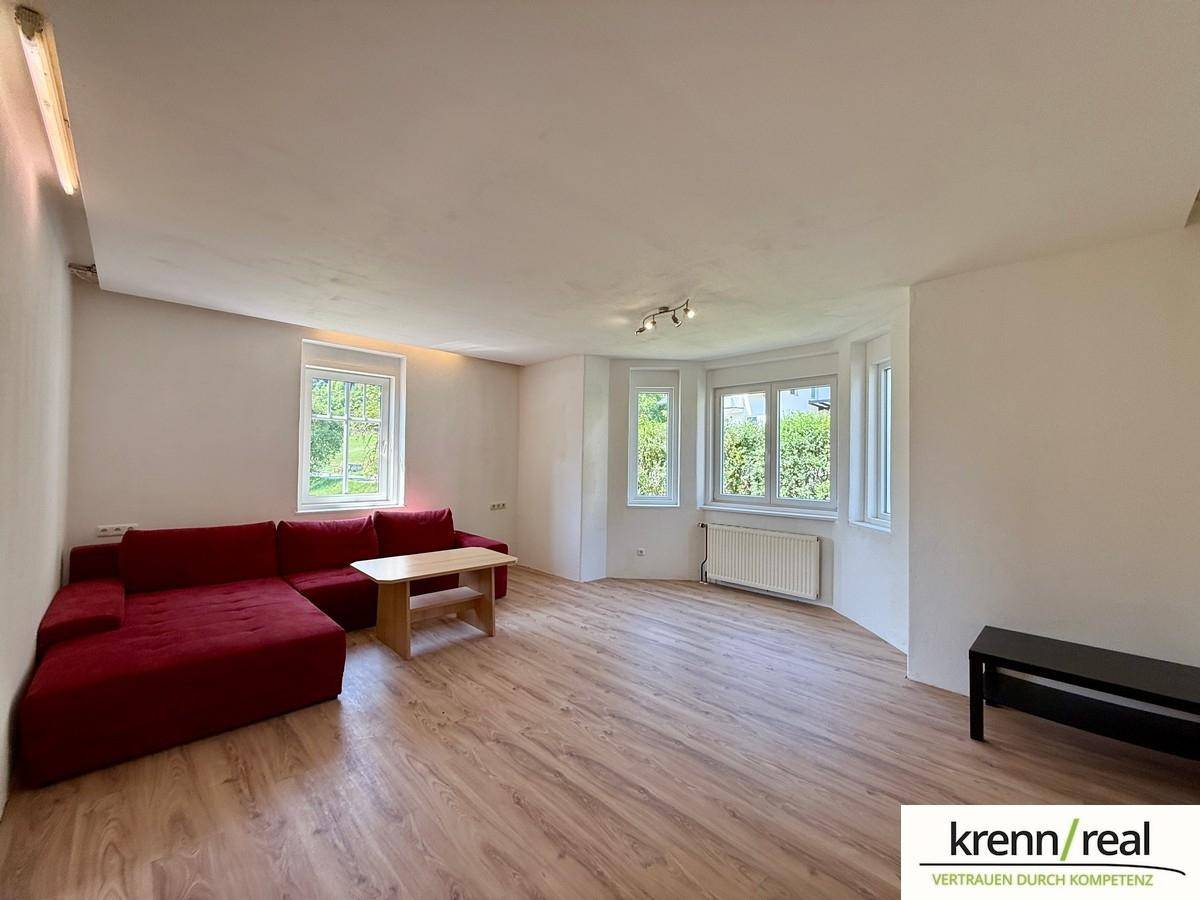 Geräumiges Wohnzimmer mit Laminatboden, rotem Sofa und Erkerfenstern mit Blick ins Grüne.