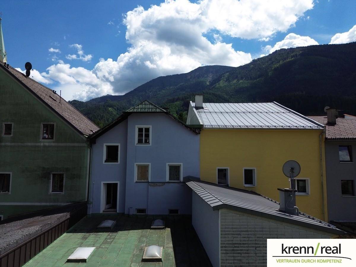 Rückansicht des hellblauen Gebäudes und eines gelben Nachbarhauses mit Bergen im Hintergrund unter blauem Himmel.