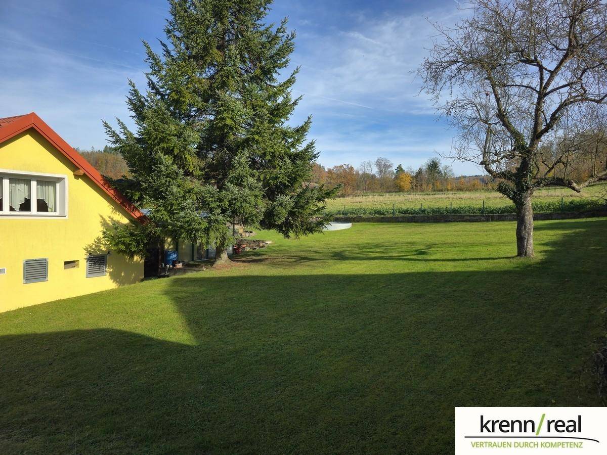 Weitläufige Rasenfläche neben einem gelben Haus mit Blick auf Felder und Wald im Hintergrund.