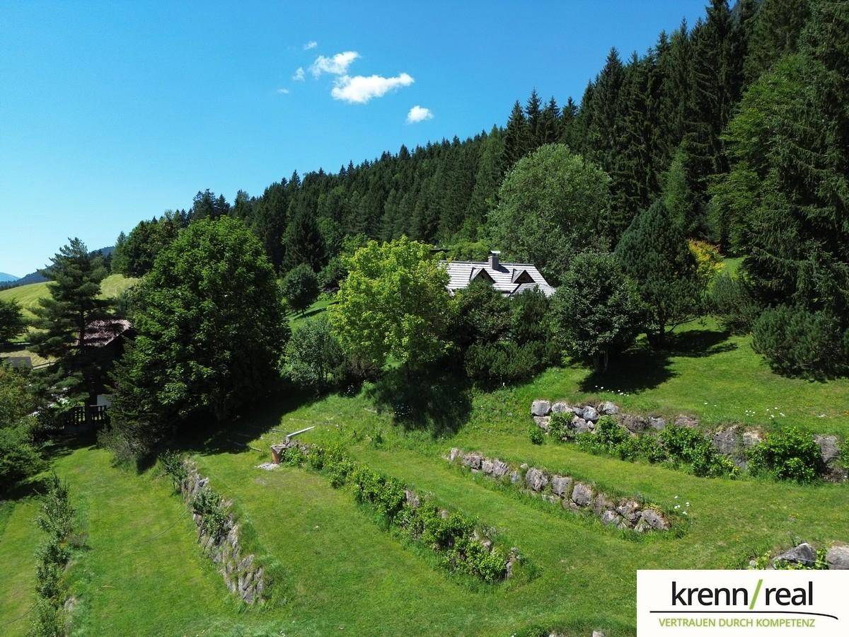 Idyllisches Haus in grüner Hügellandschaft mit dichtem Wald und terrassierten Gärten unter blauem Himmel.