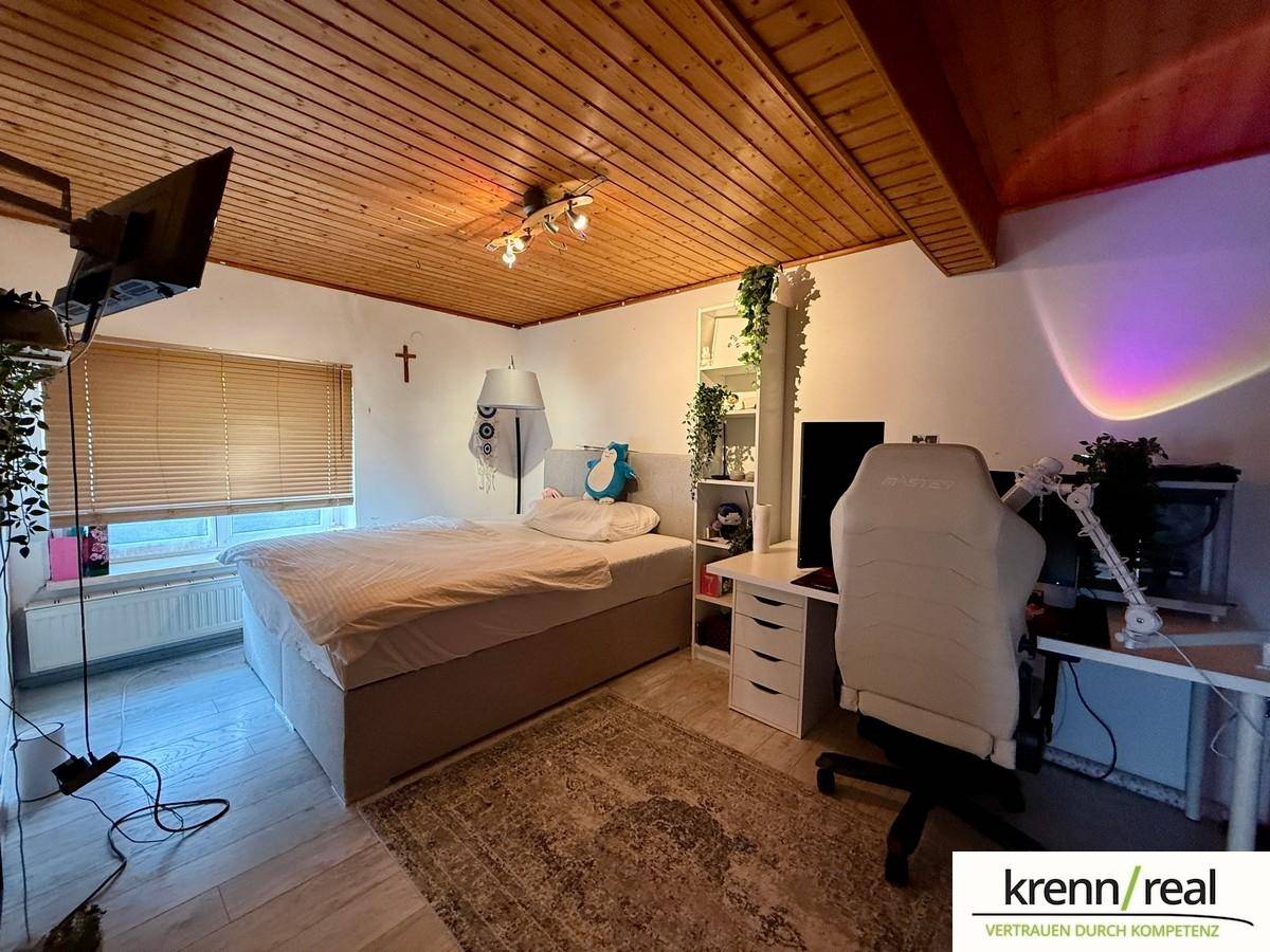 Gemütliches Schlafzimmer mit Holzbalkendecke, Bett und Arbeitsbereich, hell durch das Fenster.