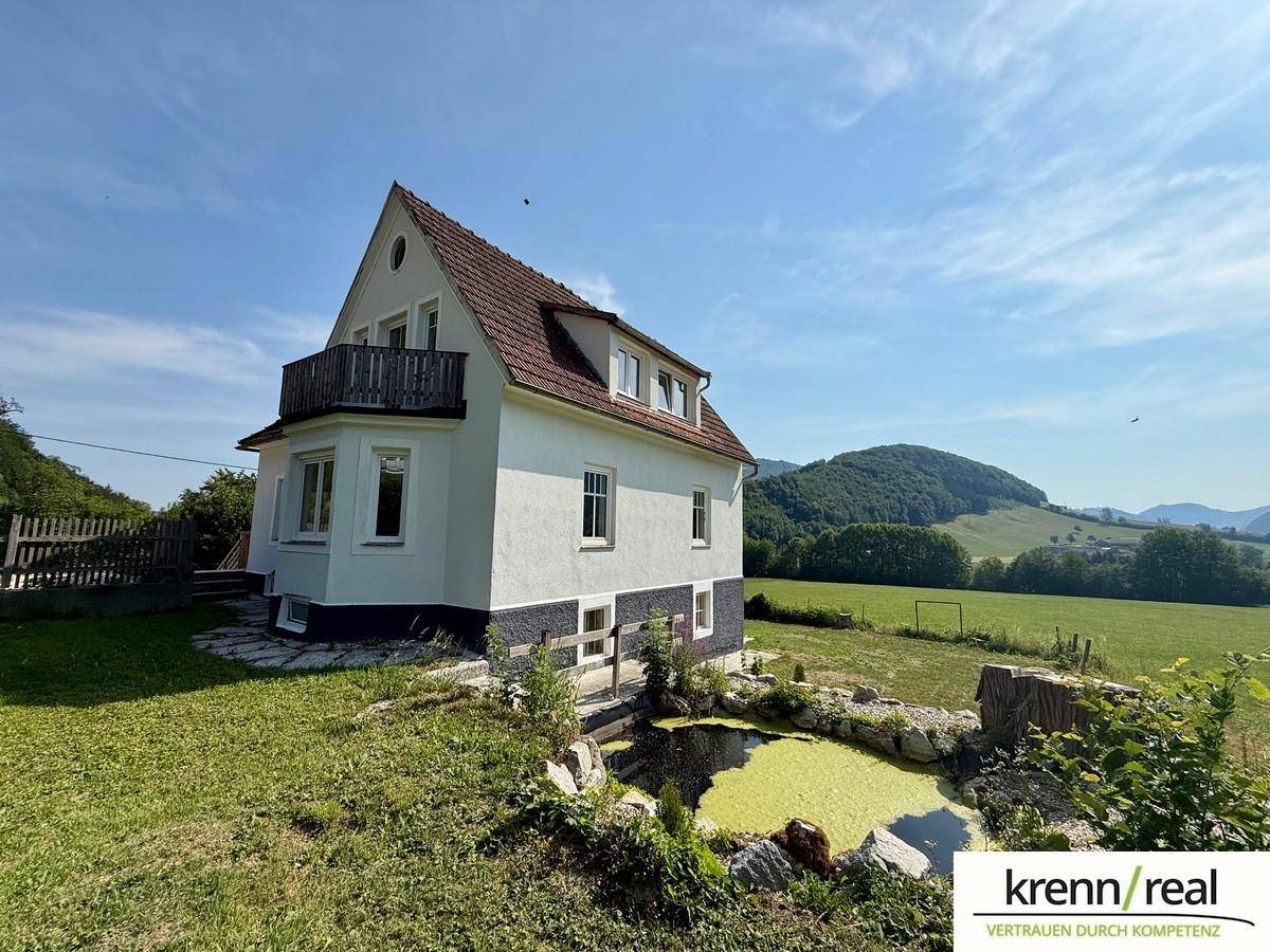 Seitenansicht des Hauses mit einem kleinen Teich im Vordergrund und einem Balkon, umgeben von grünen Feldern.