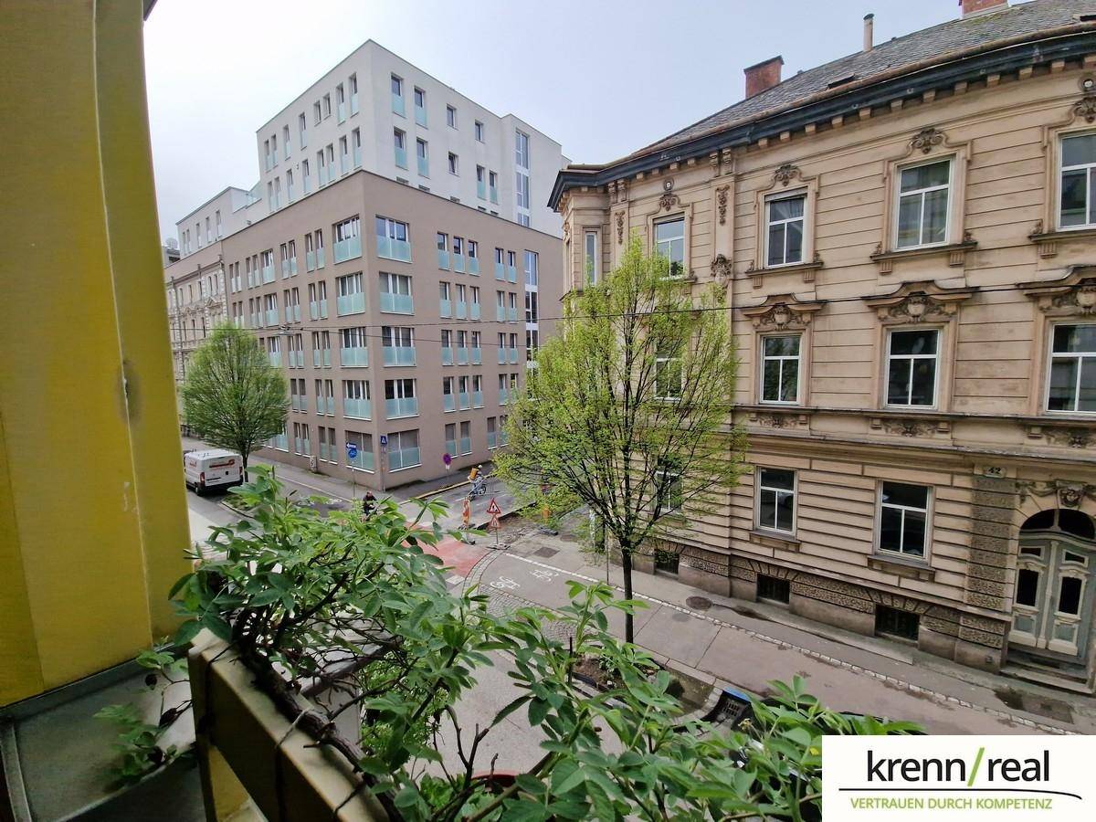 Balkon mit Blick auf die Straße, historische und moderne Gebäude sowie grüne Bepflanzung.