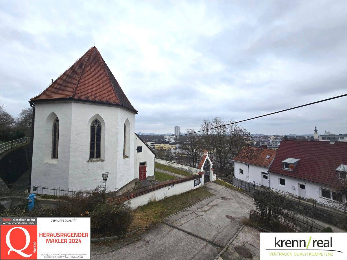 Historisches weißes Gebäude mit rotem Ziegeldach und umliegenden Häusern unter bewölktem Himmel.
