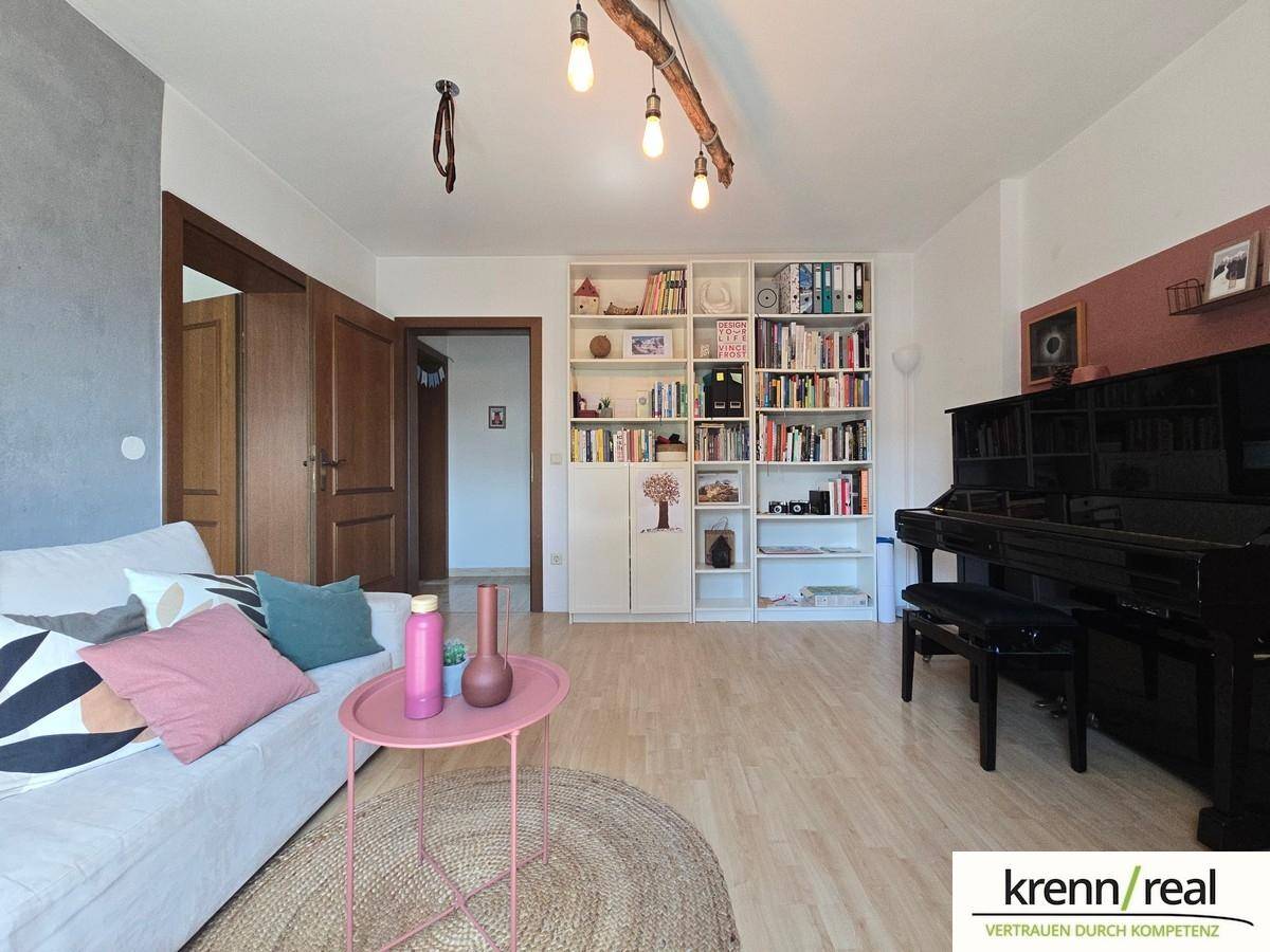 Gemütliches Wohnzimmer mit hellem Laminatboden, Bücherregal und einem schwarzen Klavier.