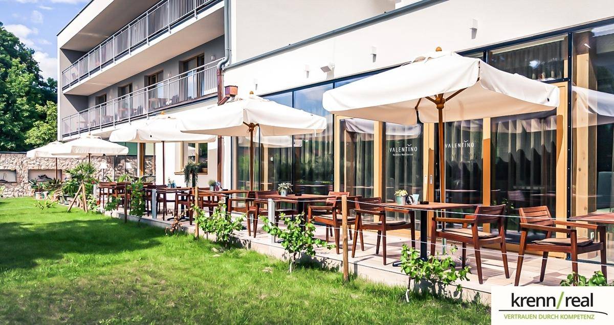 Moderne Gebäudefassade mit großen Glasfronten und einer einladenden Außenterrasse mit Sonnenschirmen und Sitzgelegenheiten.