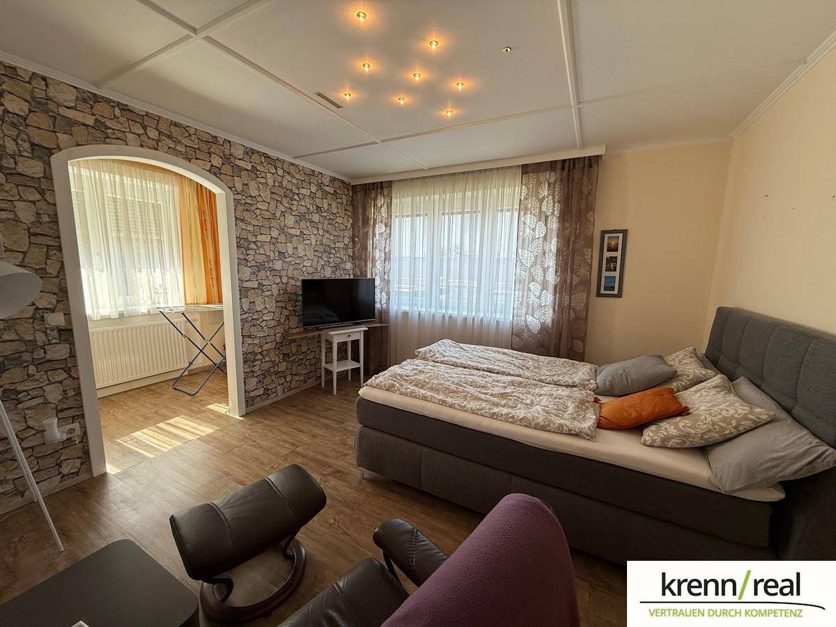 Schlafzimmer mit Bett, Akzentwand in Steinoptik und Fenster mit Vorhängen, hell beleuchtet.