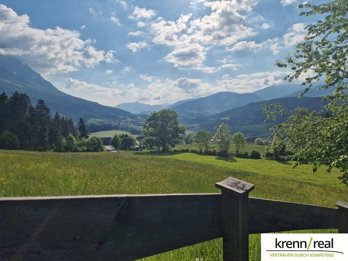 Panoramablick über ein weites Tal mit grünen Wiesen und Bergen im Hintergrund.