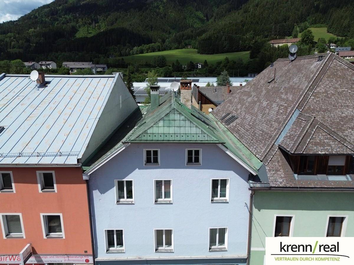 Vorderansicht des hellblauen Gebäudes mit angrenzenden Häusern, Dächern und einer Bergkulisse unter bewölktem Himmel.