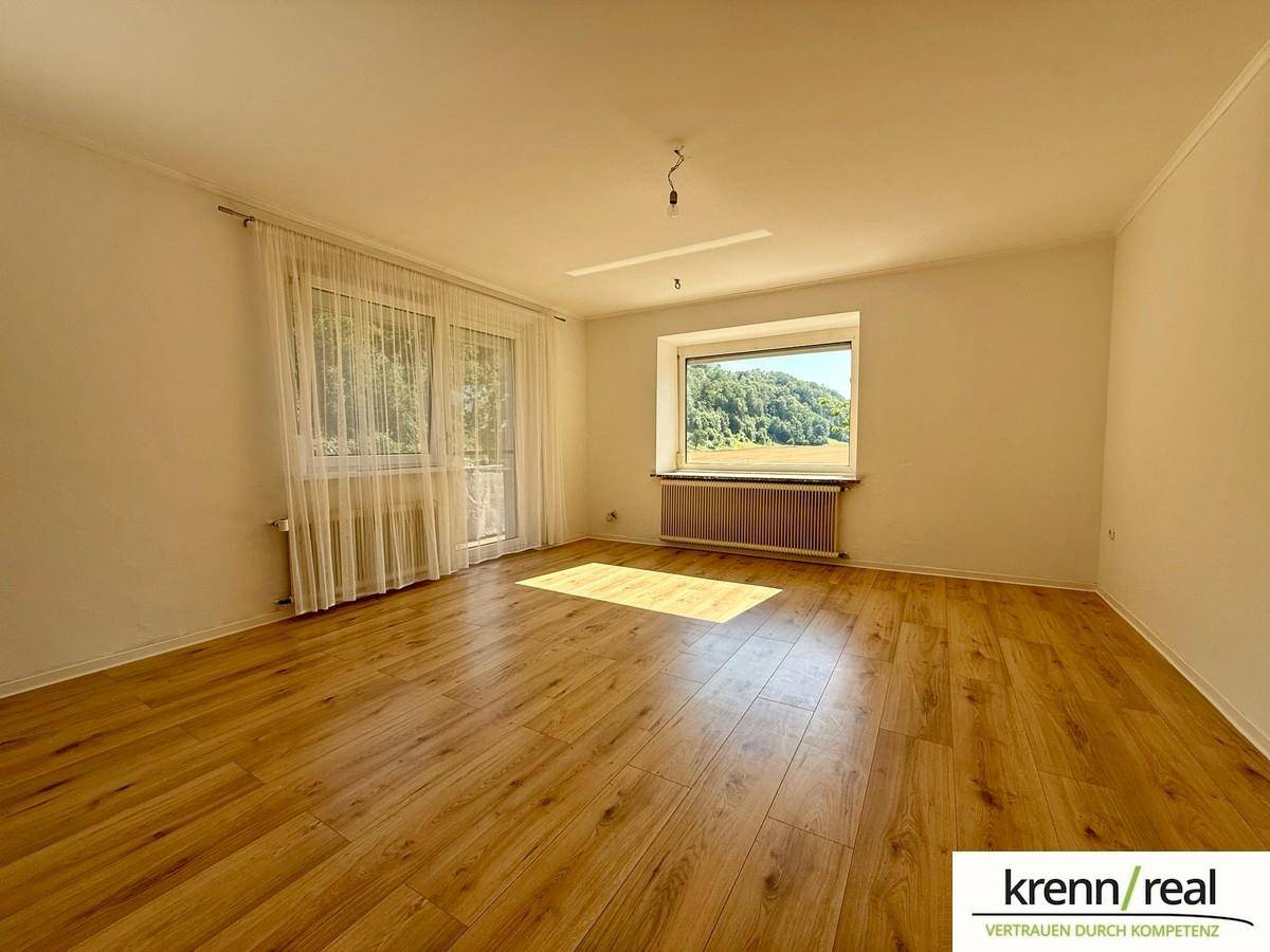 Heller Wohnraum mit Laminatboden und großen Fenstern, die viel Sonnenlicht hereinlassen.
