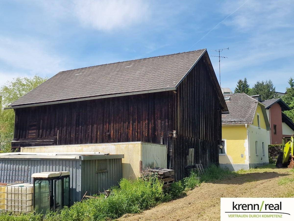 Seitenansicht der dunklen Holzscheune mit angrenzendem gelben Wohnhaus und Nebengebäuden.