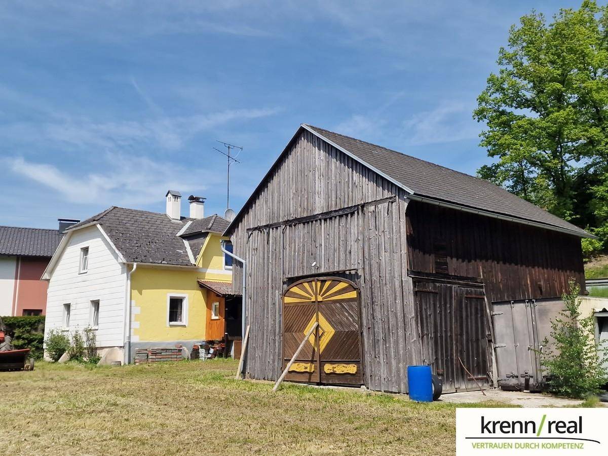 Rustikale Holzscheune mit gelbem Wohnhaus im Hintergrund unter klarem Himmel.