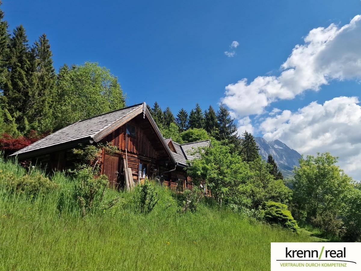 Rustikales Holzhaus in alpiner Landschaft mit grünen Wiesen und bewaldeten Bergen unter blauem Himmel.