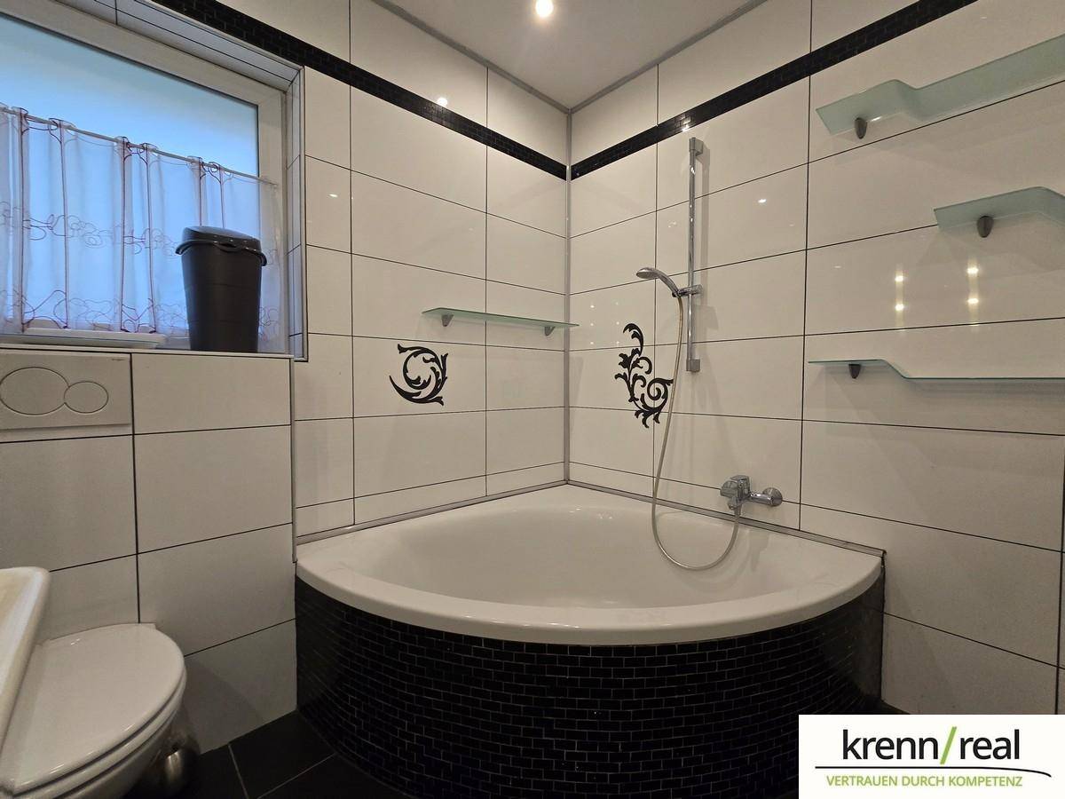 Badezimmer mit Eckbadewanne, Duschvorrichtung, Toilette und einem Fenster, gefliest in Weiß und Schwarz.