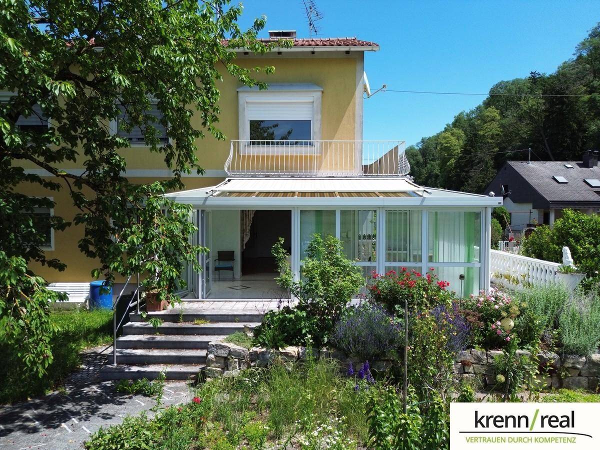 Eingangsbereich des Hauses mit Wintergarten, umgeben von einem blühenden Garten und alten Bäumen.