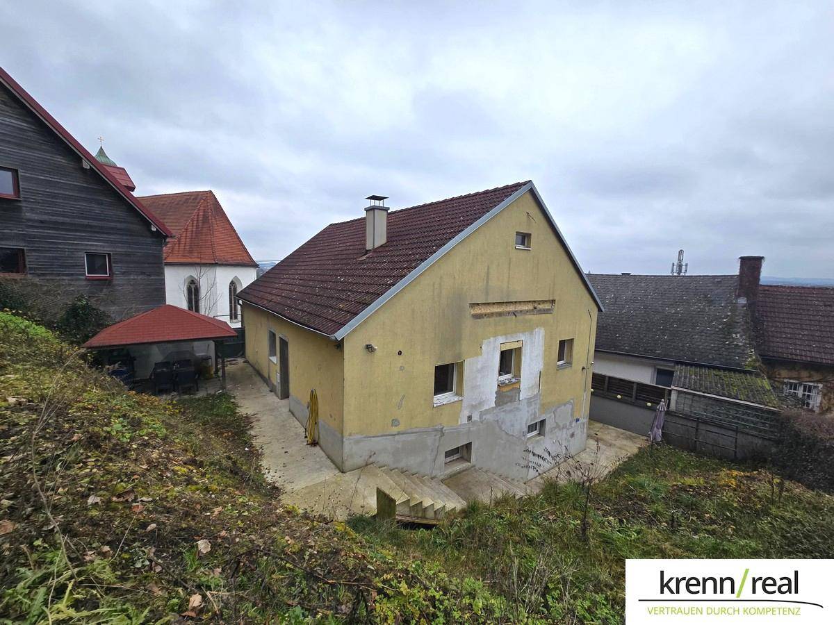 Gelbes Haus mit rotem Dach, das Renovierungsbedarf zeigt, umgeben von Vegetation und weiteren Gebäuden.