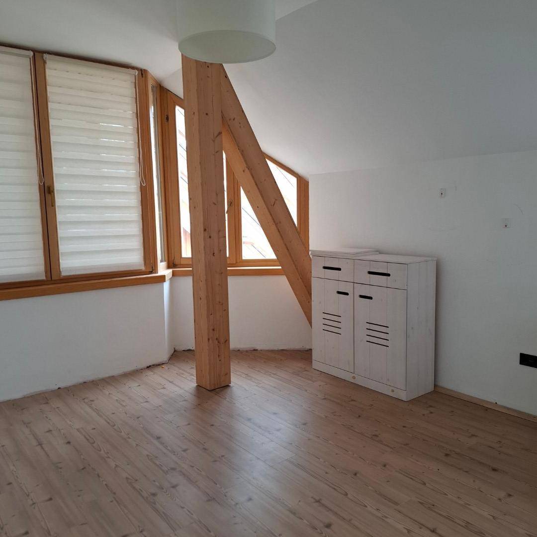 Heller Wohnraum mit Holzdielenboden, sichtbaren Holzbalken und großen Fenstern mit Jalousien unter Dachschrägen.