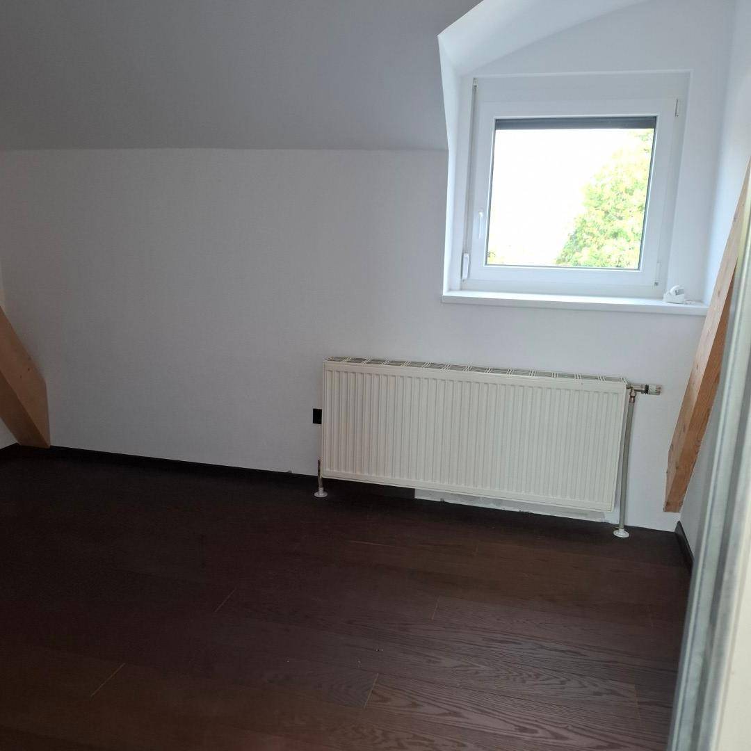 Schlafzimmer mit dunklem Holzboden, weißen Wänden, einem Fenster und Heizkörper unter einer Dachschräge.