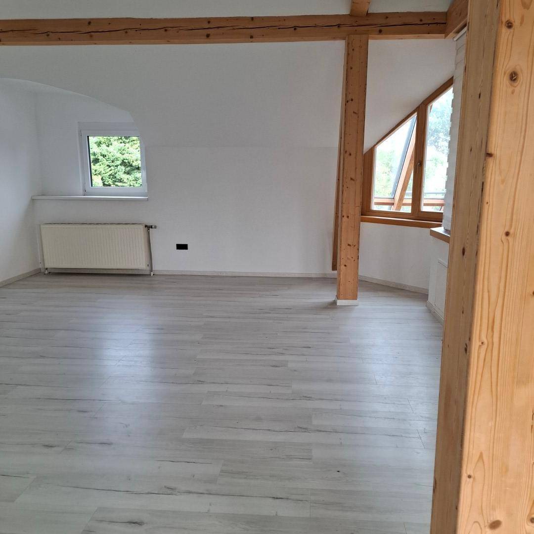 Geräumiger Wohnraum mit hellem Laminatboden, weißen Wänden, sichtbaren Holzbalken und Fenster mit Heizkörper.
