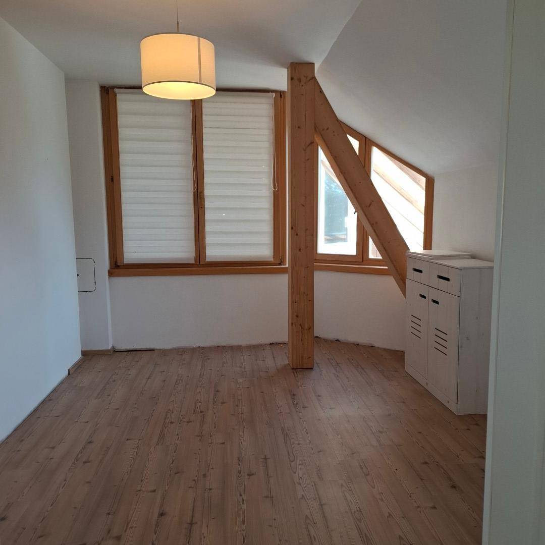 Geräumiges Zimmer im Dachgeschoss mit Holzbalken, Dachschräge und mehreren Fenstern.